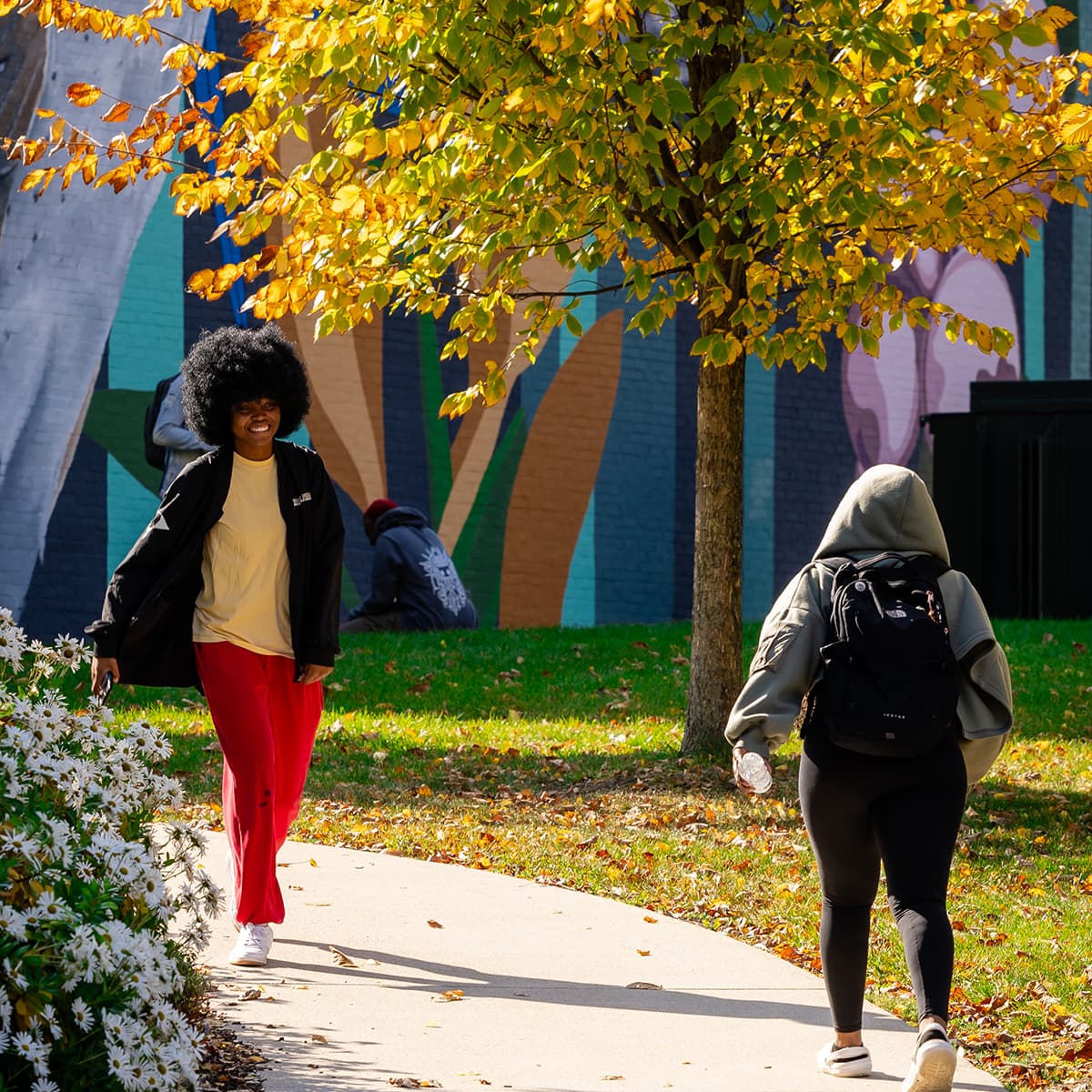 Two students walking past a mural on a sunny fall day, one smiling under golden leaves.