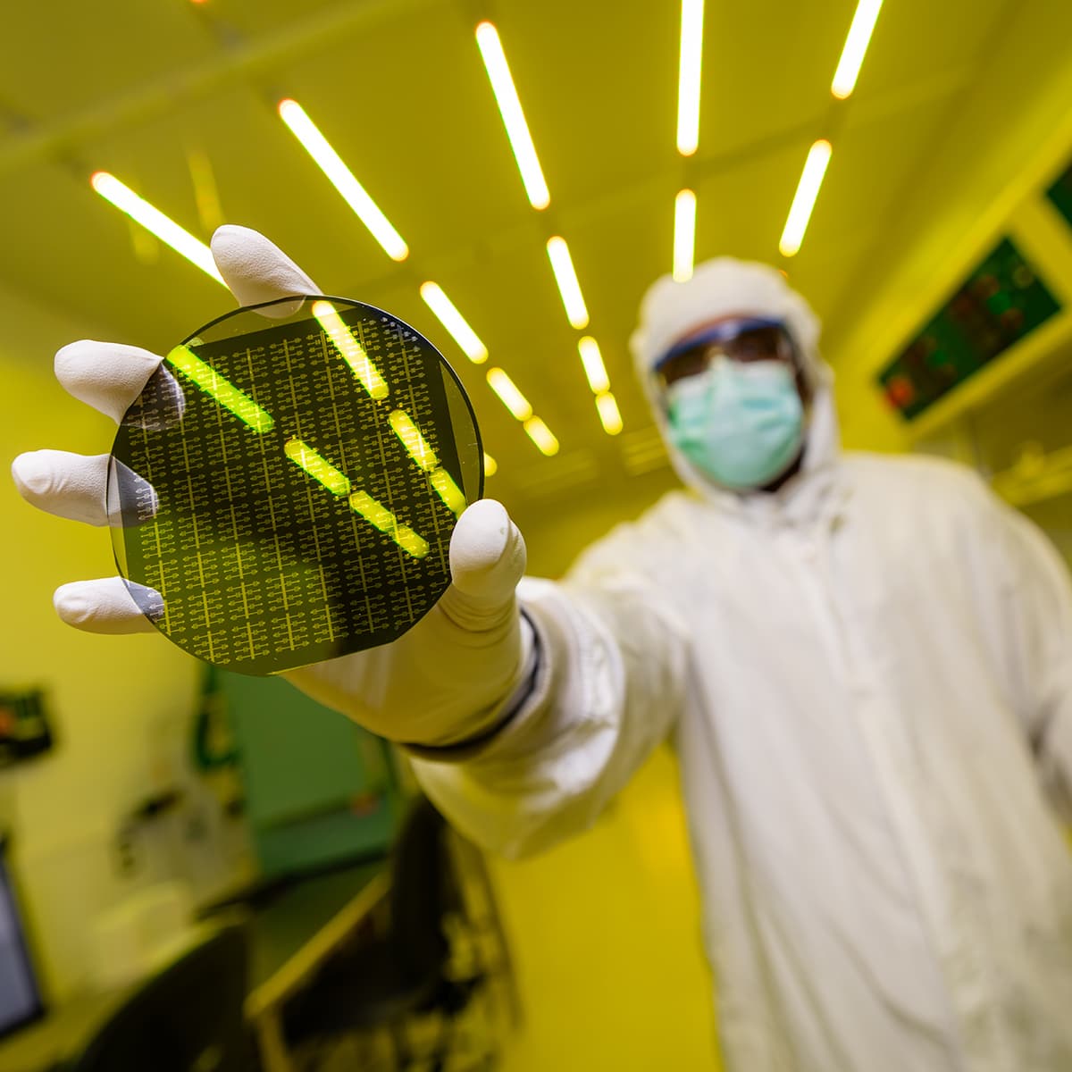 Scientist in cleanroom suit holding a silicon wafer in a lab with yellow lighting