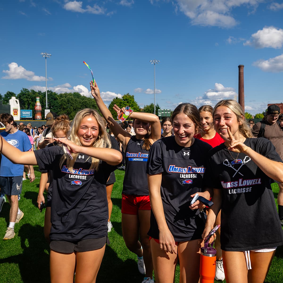 Group of UMass Lowell lacrosse players smiling and celebrating outdoors on a sunny day.