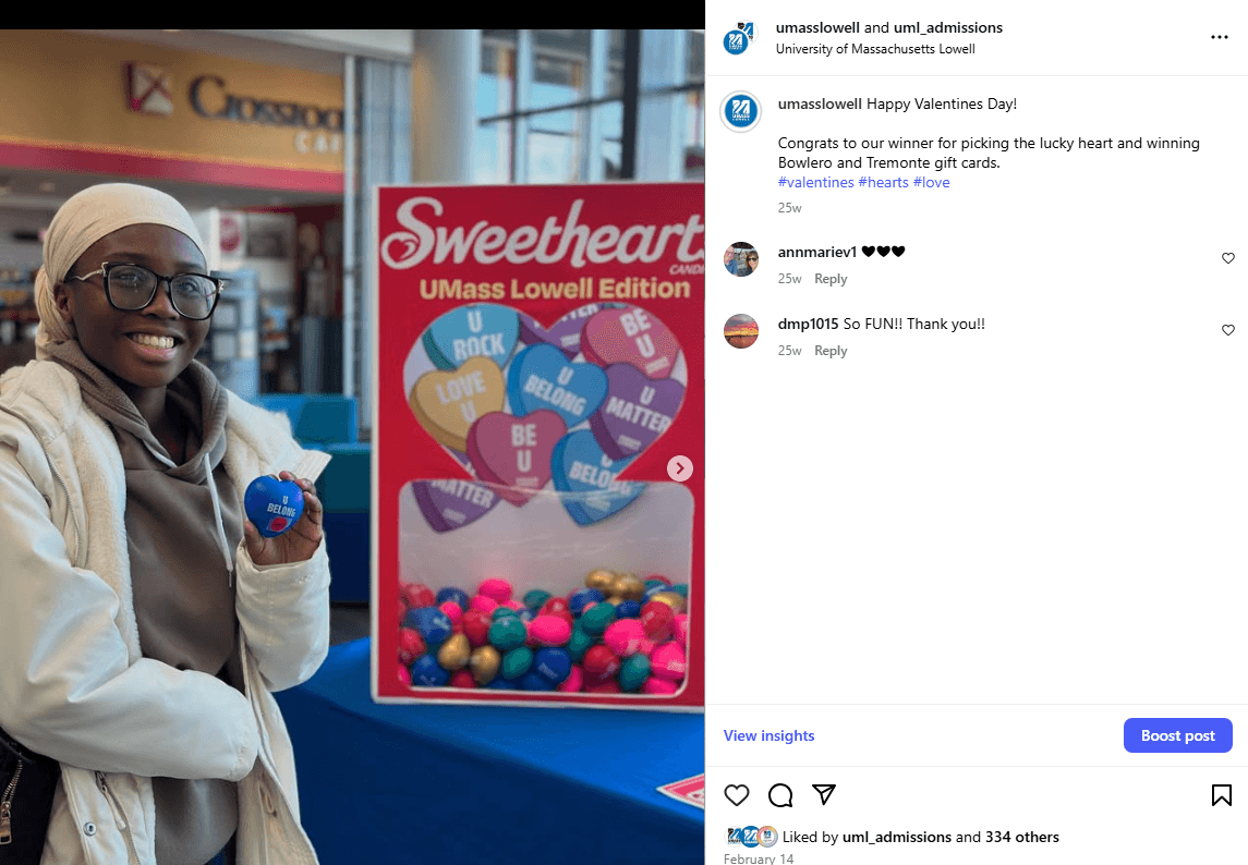 Student smiling and holding a “U Belong” Valentine heart in front of UMass Lowell Sweetheart display.